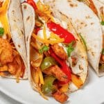 Trois tortillas garnies de poulet, poivrons rouges et verts, crème sure et fromage râpé sur une assiette blanche.
