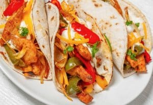 Trois tortillas garnies de poulet, poivrons rouges et verts, crème sure et fromage râpé sur une assiette blanche.