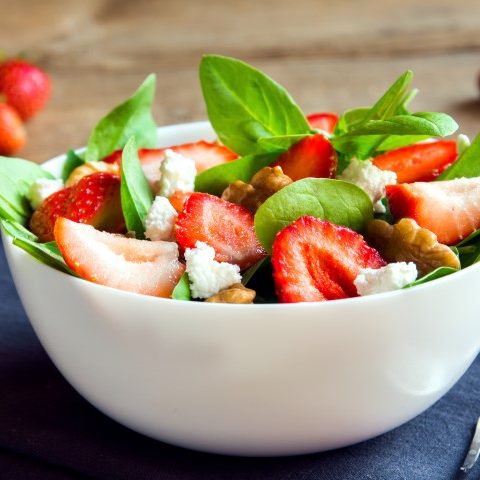 Salade composée de bébés épinards, de fraises, pacanes rôties, fromage de chèvre avec une vinaigrette légèrement sucrée au miel servie dans un bol blanc.
