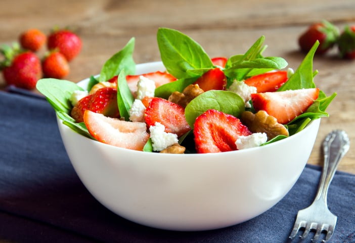 Salade composée de bébés épinards, de fraises, pacanes rôties, fromage de chèvre avec une vinaigrette légèrement sucrée au miel servie dans un bol blanc.